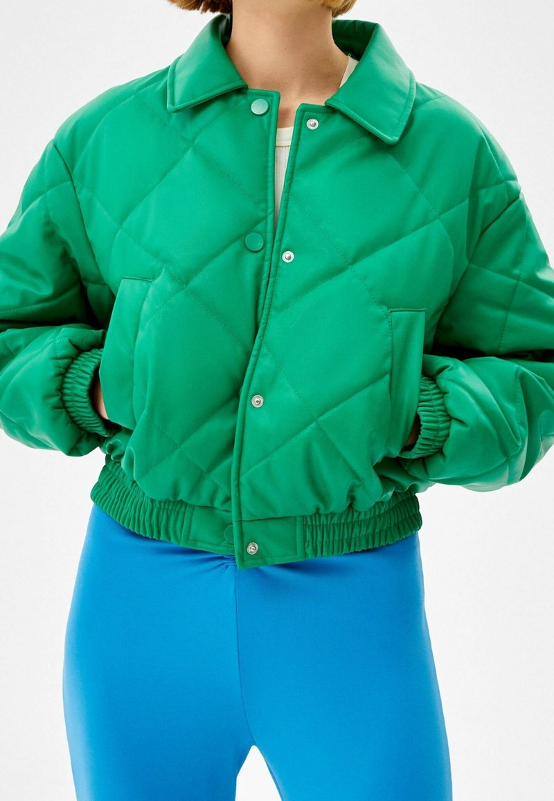 Zelená prošívaná zkrácená bunda s vysokým límcem, patentkami a elastickými manžetami. Kombinováno s modrými úzkými legínami.