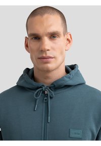 Felpa con zip verde acqua e collo con cordino, caratterizzata da un tessuto texturizzato e un'etichetta del marchio sul lato sinistro. Superficie liscia, design casual.