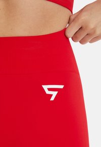 Leggings de sport rouges à côtes avec un logo blanc sur la taille. Le matériau semble extensible, conçu pour le confort et le mouvement.