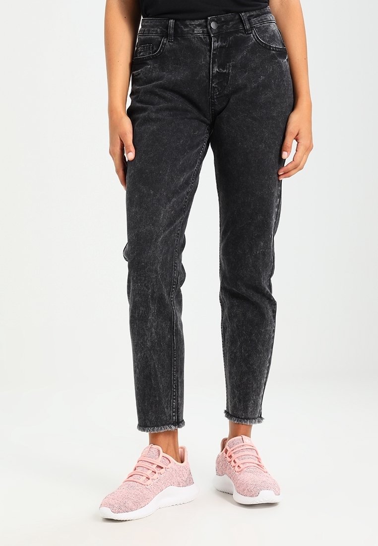 Mustat, korkeavyötäröiset farkut himmeällä tekstuurilla, viisi taskua ja käännetyt lahkeet, yhdistetty vaaleanpunaisiin neulottuihin tennareihin.