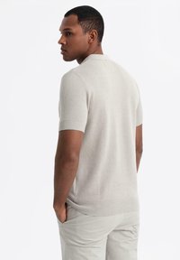 Ljusbeige kortärmad polo med en strukturerad stickad mönster, ribbad krage och ärmslut; bärs med ljusa byxor, händerna i fickorna.