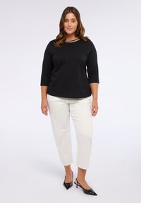 Mujer de pie con un top negro con detalles de cadena, pantalones cortos blancos y zapatos de tacón destalonados negros de punta en un fondo blanco.