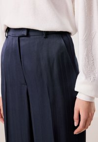 Marineblaue, gefaltete Hose mit Fischgrätenmuster, ausgestattet mit einem tiefen Bund und glatter Textur, kombiniert mit einem weißen Pullover mit Perlendetails.