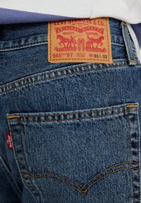 Jeans de ganga azul-escuro com um padrão distintivo de costura laranja. Apresenta uma etiqueta de couro castanho com logótipo em relevo e detalhes do modelo.