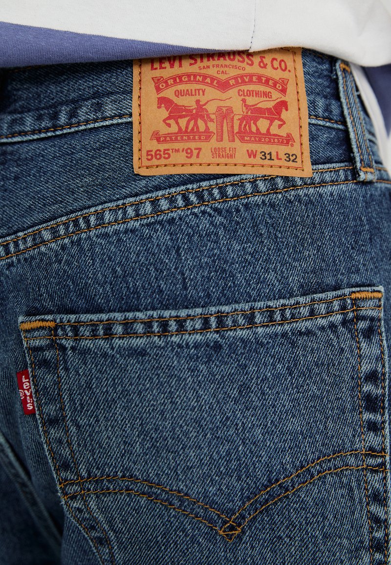 Jean en denim bleu foncé avec un motif de couture orange distinctif. Inclut une étiquette en cuir marron avec un logo embossé et des détails sur le modèle.