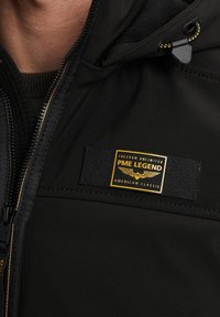 Zwarte geïsoleerde jas met een gladde textuur, voorzien van een opvallende gele en zwarte merkembleem en ritsdetails.