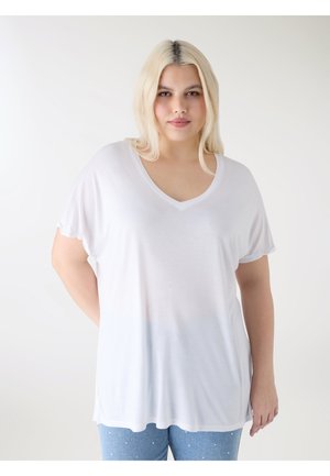 Blondynka stojąca z rękami za plecami, ubrana w luźną białą koszulkę z dekoltem w serek i jasnoniebieskie dżinsy w kropki, neutralne tło.