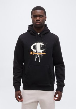 Homme portant un sweat à capuche noir avec le logo Champion blanc et orange dégoulinant, associé à un pantalon de couleur claire, debout devant un fond uni.