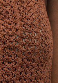 Κοντινό πλάνο καφέ πλεκτού υφάσματος με μοτίβα διαμαντιών και τετραγώνων πάνω από ανοιχτό μπλε ένδυμα, με ορατό σκούρο δέρμα στο πλάι.