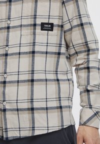 Camicia button-up in beige chiaro con un motivo a quadri blu navy e beige. Presenta una tasca sul petto con un'etichetta nera e bottoni bianchi. Maniche lunghe.