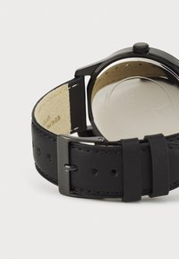 Crni kožni remen za sat s teksturiranim finišem, siva metalna kopča i glatka metalna stražnja strana, koja prikazuje minimalistički dizajn.