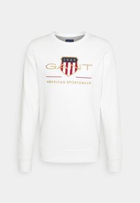 Weißes Sweatshirt mit gerippten Bündchen und Saum, ausgestattet mit einem goldenen und roten gestickten Logo, das die Schriftzüge "GANT" und "AMERICAN SPORTSWEAR" trägt.
