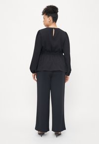 Black, long-sleeve top with a gathered waist and keyhole back detail, paired with wide-leg black trousers and black heeled shoes.