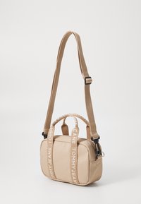 Geantă crossbody bej cu bretele din material care prezintă logo-ul "TOMMY JEANS". Formă rectangulară, închidere cu fermoar, două mânere superioare și curea ajustabilă.