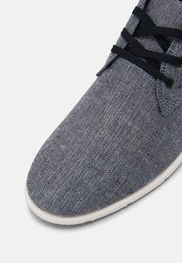 Modrá látková bota s texturovaným povrchem, kulatou špičkou a bílou gumovou podešví. Černé tkaničky jsou provlečeny očky poblíž kotníku.