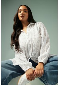 Blusa bianca e texture con maniche a sbuffo e chiusura con bottoni, abbinata a jeans blu denim e sneakers bianche. Sullo sfondo un ambiente tenue.