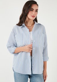 Leichtes, knöpfbares Hemd in Weiß mit blauen vertikalen Streifen. Verfügt über einen Kragen, zwei Brusttaschen und hochgekrempelte Ärmel.