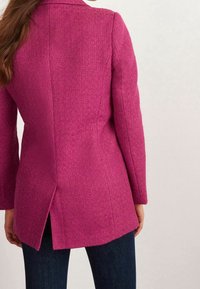 Fuksjowy płaszcz dopasowany z teksturowanej tkaniny, z długimi rękawami, klapami kołnierza i rozcięciem z tyłu dla łatwości ruchów. Nosi się go z ciemnymi dżinsami.