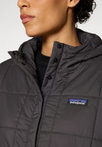 Zwarte gewatteerde jas met capuchon, drukknoopsluiting en een Patagonia-logo op de borst, gemaakt van soepel, duurzaam materiaal.