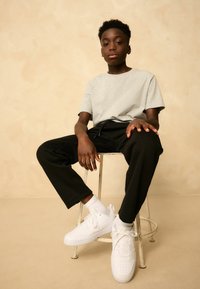 Graues kurzärmliges T-Shirt, schwarze schmal zulaufende Hose und weiße Sneakers, getragen mit weißen Socken. Die Person sitzt auf einem abgenutzten Metallhocker.