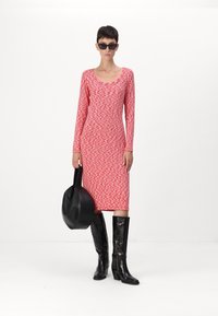 Enganliegendes Langarmkleid mit einem rot-weißen Wellenmuster, kombiniert mit schwarzen knielangen Stiefeln und einer geschwungenen schwarzen Tragetasche.