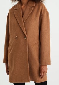 Brązowy dwurzędowy płaszcz z mieszanki wełny z wyciętym kołnierzem, dwoma przednimi kieszeniami i ozdobnymi guzikami. Miękka faktura i luźny krój.