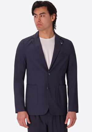 Uomo che indossa una giacca da completo blu scuro e pantaloni abbinati sopra una camicia bianca, che guarda di lato su sfondo semplice.