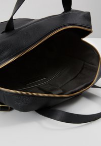 Zwarte leren handtas open om een ruime binnenkant met stoffen voering en gouden rits te tonen, rustend op een witte ondergrond.