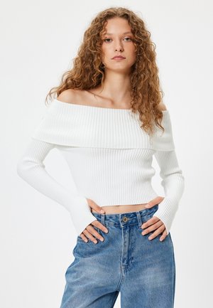 Witte geribbelde off-the-shoulder crop top met lange mouwen, gedragen met lichtblauwe high-waisted jeans, die een strak textuur en ontwerp tonen.