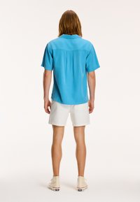 Lyseblå kortærmet skjorte med en rygflap, knapkrave og sidespidser, parret med hvide shorts og beige sneakers.