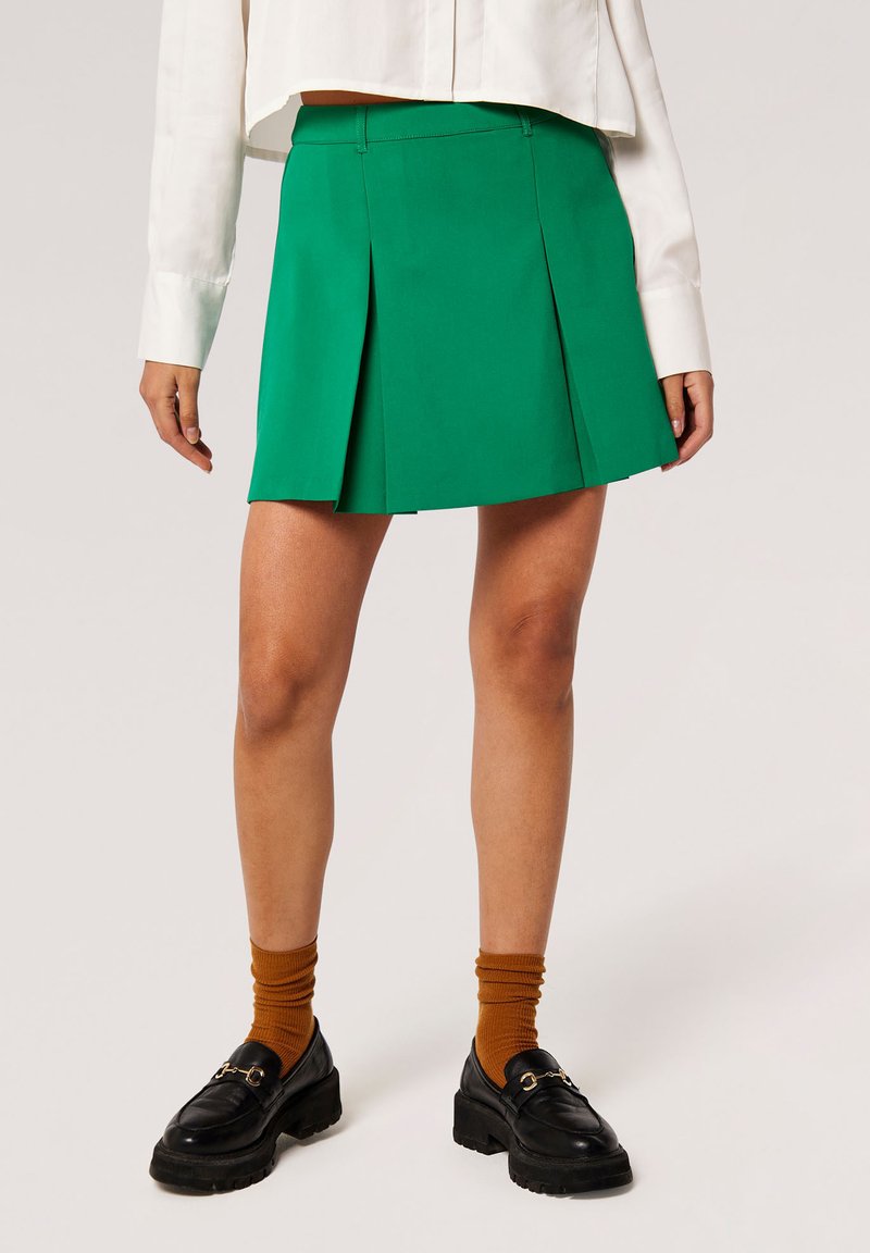 Grüne, plissierte Minirock aus glattem Stoff mit einer mittelhohen Taille. Kombiniert mit schwarzen Slippern und braunen, gerippten Socken.