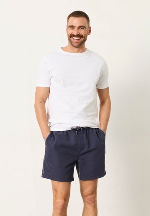 Mann mit kurzen Haaren und Schnurrbart, der ein weißes T-Shirt und navyblaue Shorts trägt, steht mit den Händen in den Taschen und lächelt vor einfarbigem Hintergrund.