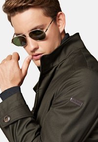 Oliwkowa kurtka z wysokim kołnierzem, guzikami i markową naszywką na rękawie. Model nosi okulary przeciwsłoneczne i dotyka ust.