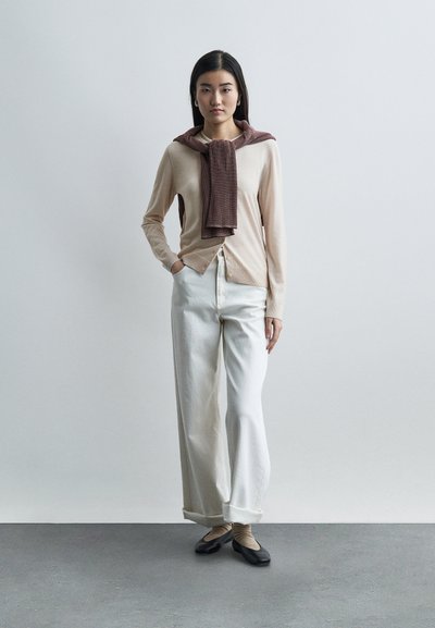 Γυναίκα όρθια με τα χέρια στις τσέπες, φοράει μπεζ ζακέτα, καφέ πουλόβερ περασμένο στους ώμους, άσπρο παντελόνι φαρδιάς γραμμής, μπεζ κάλτσες και μαύρα μπαλαρίνες.