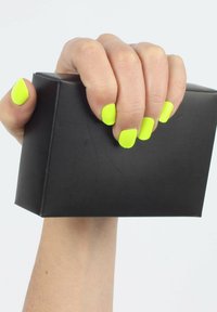 Hand, die einen schwarzen rechteckigen Block mit leuchtend neon-gelben lackierten Nägeln vor einem schlichten hellen Hintergrund hält.