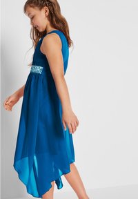 Un vestito senza maniche di un blu brillante, realizzato in tessuto leggero e trasparente, con una cintura decorativa a motivi e un orlo posteriore più lungo.