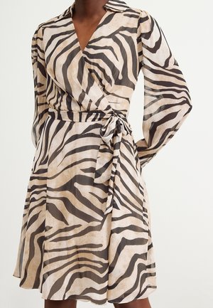 Vrouw draagt een beige-zwart zebraprint wikkeljurk met lange mouwen en een zijstrik in de taille.