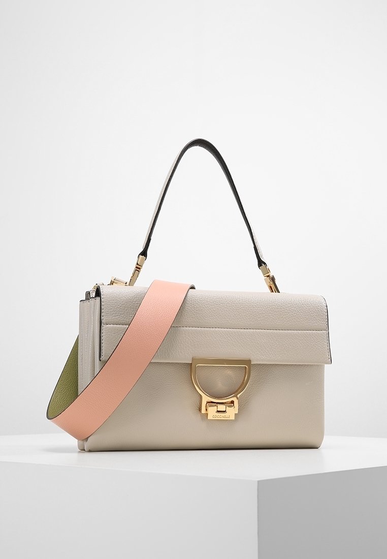 Sac à main en cuir beige avec une bandoulière rose et verte, une poignée supérieure noire et un fermoir en métal doré sur un socle blanc.