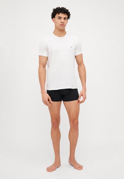 Tricou alb cu mânecă scurtă, cu guler rotund și un logo mic pe piept, asortat cu pantaloni scurți negri, ajustați. Modelul stă desculț.