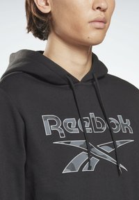 Sudadera negra con cordón, que presenta un logo de Reebok gris y acentos de camuflaje. Tejido suave con un corte regular y puños acanalados.