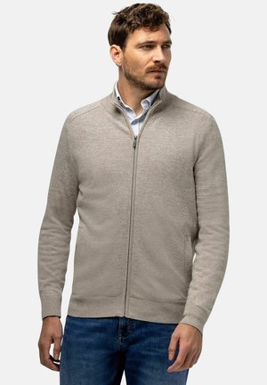 Man met kort bruin haar en baard, die een beige vest met rits draagt over een lichtblauw shirt en blauwe spijkerbroek, kijkt naar rechts met een neutrale uitdrukking.