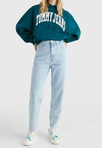 Felpa con cappuccio color teal con la scritta "TOMMY JEANS" stampata in bianco, jeans azzurri chiari ricamati e sneakers multicolori con un design texturizzato.