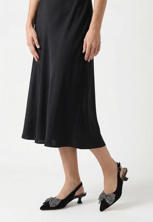 Frau trägt einen schwarzen Midirock und schwarze Slingback-Schuhe mit kleinem Absatz und großen Strassschleifen.