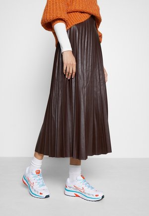 Person trägt einen braunen plissierten Midirock, einen orangen Strickpullover, ein weißes Langarmshirt, weiße Socken und weiße Sneakers mit orangefarbenen und blauen Akzenten.