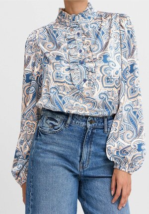 Femme portant un chemisier à manches longues à motifs cachemire avec volants et un jean bleu taille haute, debout avec une main détendue sur le côté.