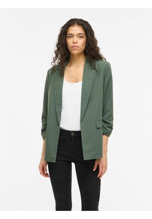 Kobieta o kręconych włosach, ubrana w zielony żakiet na białej bluzce i czarnych spodniach, stojąca na tle jednolitego białego tła.