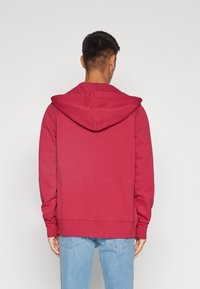 Sudadera roja con capucha de corte holgado, con bolsillo tipo canguro y puños acanalados, combinada con vaqueros azul claro.