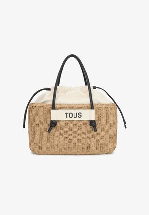 Sac en paille tissé avec des poignées en cuir noir, un haut à cordon crème, et une bande blanche avec le texte "TOUS." Design rectangulaire texturé.