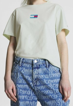 Persoon draagt een lichtgekleurde Tommy Jeans T-shirt en blauwe spijkerbroek met herhaaldelijk "Tommy Jeans" gedrukt in een groot, gestileerd lettertype.