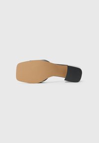 Black high-heeled shoe with a square toe and a textured rubber sole. The heel is lined with smooth black material, contrasting the tan sole.